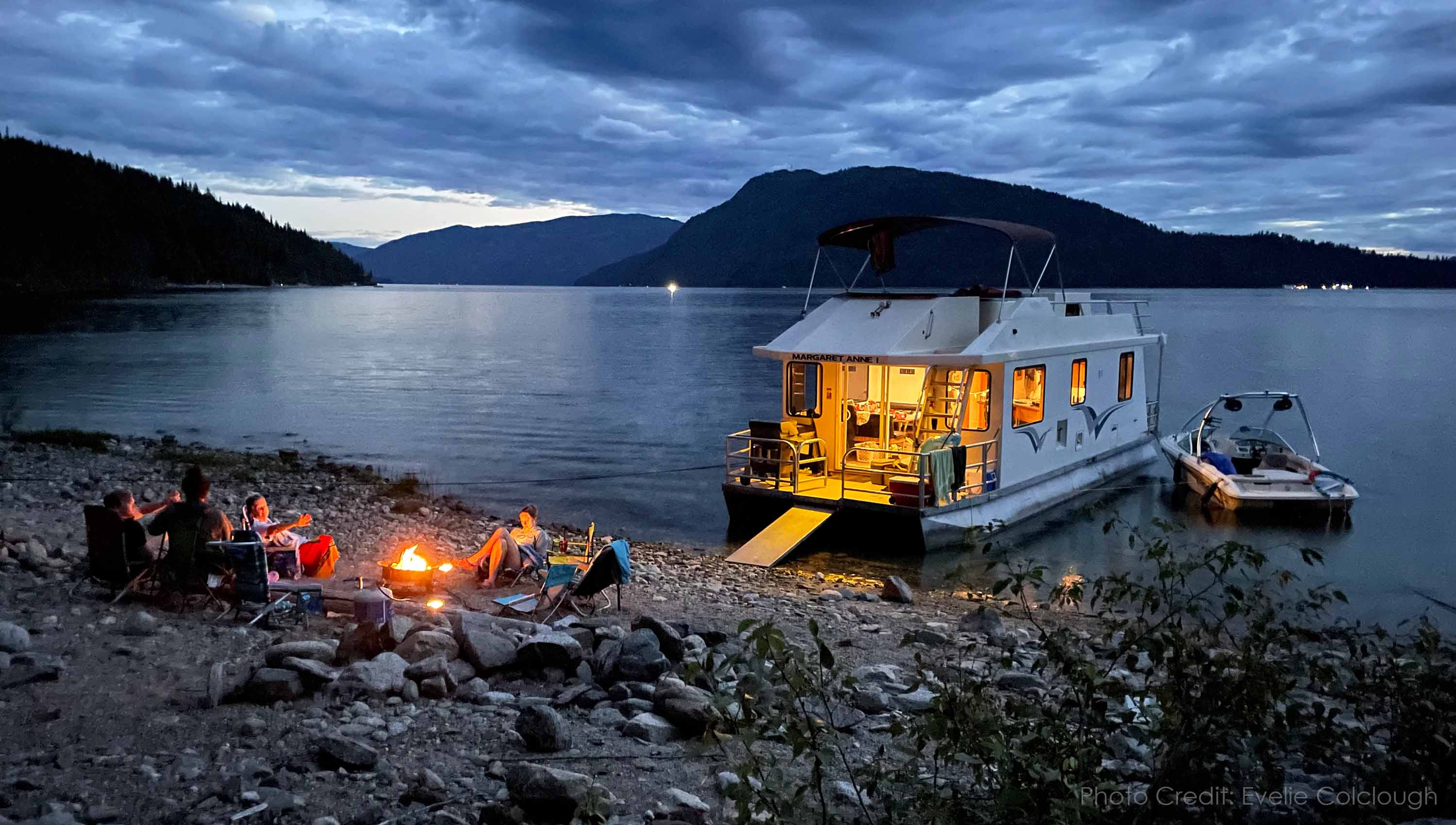 A houseboat beached at dusk with a family enjoying a campfire on the beach.