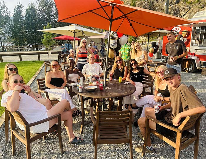 A large group enjoying food and beer on the Sausages of Sicamous eating area on a summers evening.